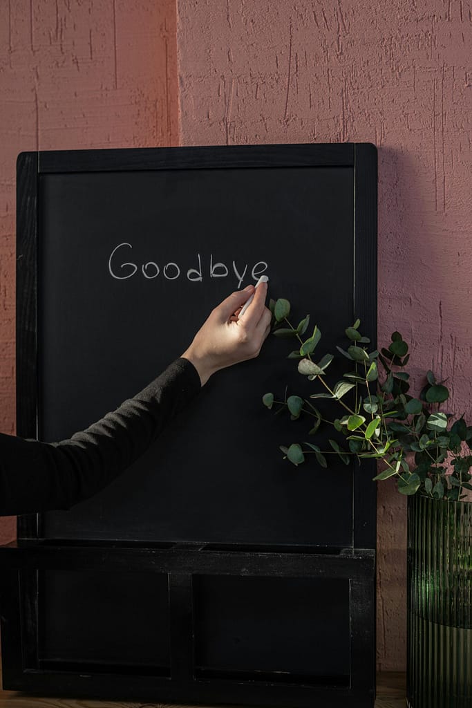 A hand writes 'Goodbye' on a chalkboard, indoors, with elegant plant decor.